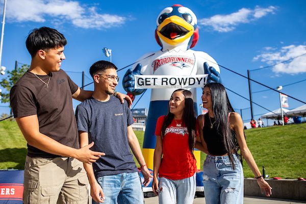 Four MSU Denver students stand smiling and laughing together in front of a large inflatable Roadrunners mascot holding a “GET ROWDY!” sign. They are outdoors on a sunny day with a grassy hill and a bright blue sky in the background, celebrating school spirit and camaraderie.