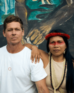 Mitch Anderson, wearing a white t-shirt, Nemonte Nenquimo wearing a yellow shirt with bead work and red paint across her eyes. Nemonte is leaning on her husband, Mitch. 