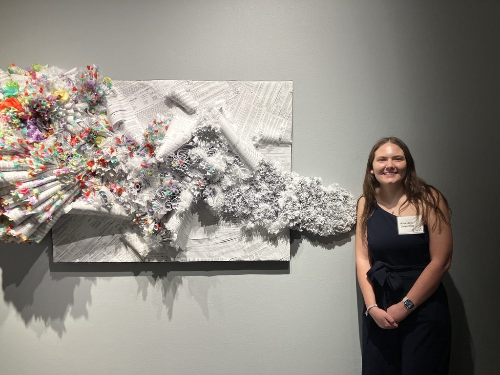 A young woman stands next to a paper sculpture mounted to an art gallery wall.