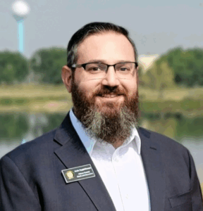 Man smiling outdoors, wearing glasses and a grey blazer. 