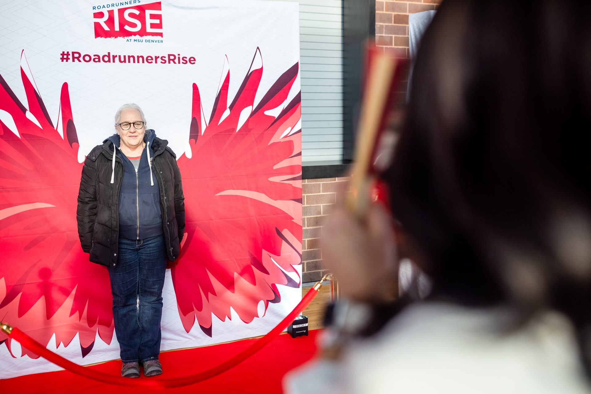 A person stands in front of a backdrop with red wings and the Roadrunners Rise logo