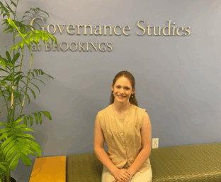 a young woman wearing a tan dress sits in front of a blue wall at the Brookings Institution, in the Governance Studies wing of the building.