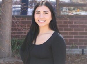 a young woman with long dark hair stands in front of a brick building, smiling.