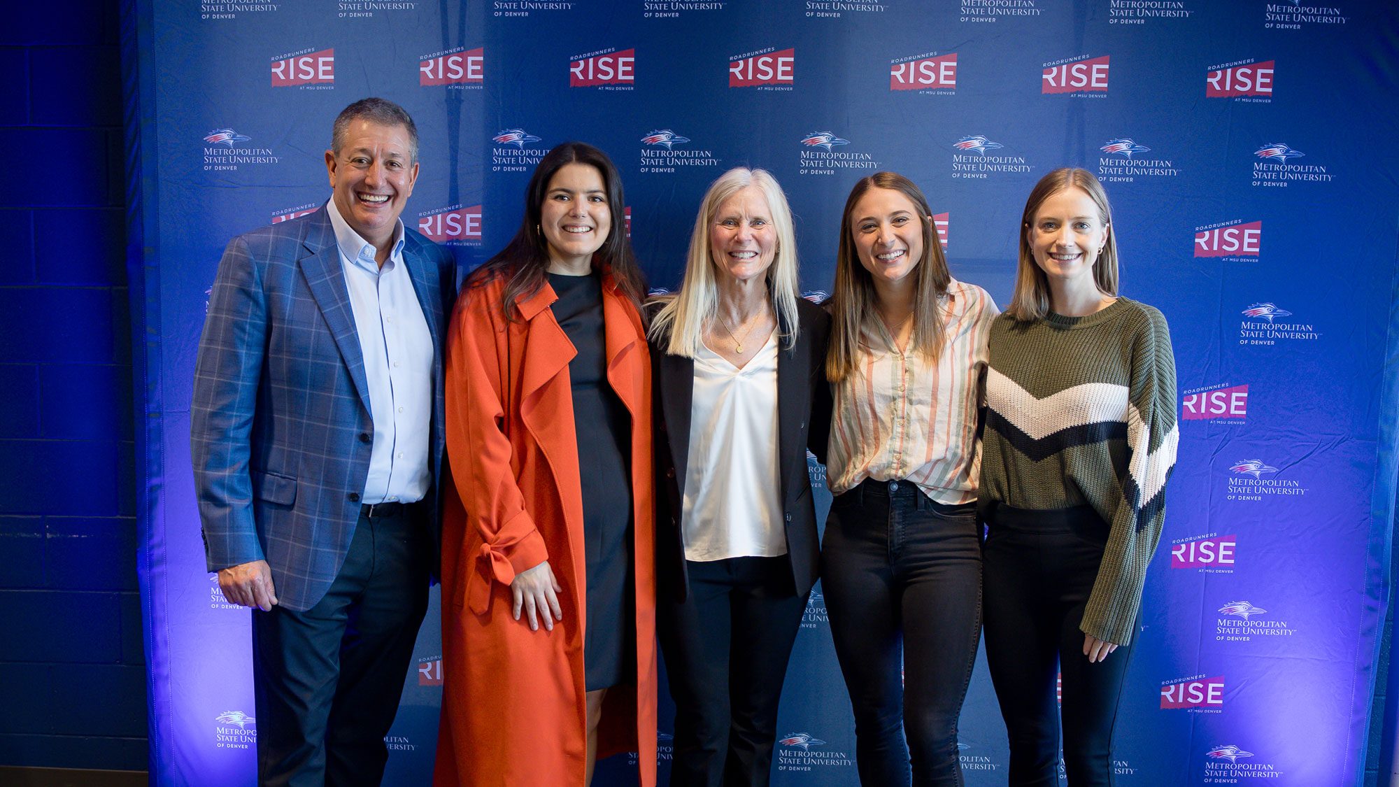 Three MSU Denver students standing for a photo with Rob and Molly Cohen in front of a Roadrunners Rise backdrop