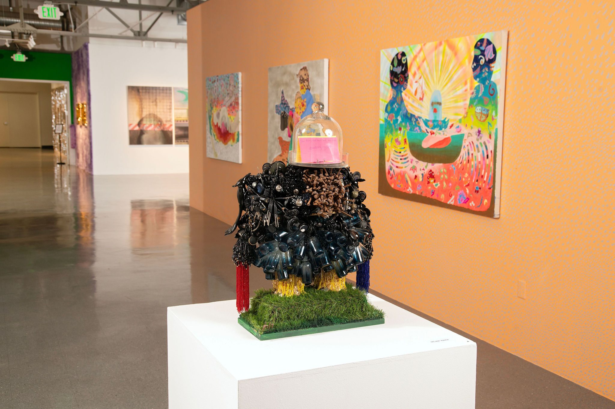 Sculpture on a pedestal in an art gallery with colorful paintings on an orange wall behind.
