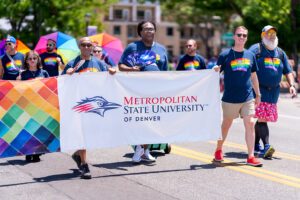 Group of people marching in a parade holding a large banner that reads ‘Metropolitan State University of Denver’ with the school’s logo. Participants are wearing matching navy blue shirts with rainbow designs and carrying colorful rainbow umbrellas. The left side of the banner features a vibrant rainbow diamond pattern. The scene takes place outdoors on a sunny day with trees and buildings in the background.