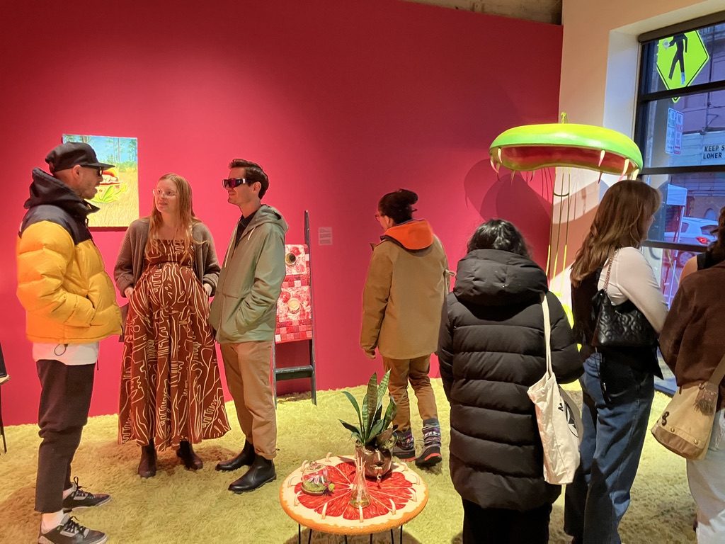A group of people stand within an art installation. The walls are painted dark pink and the floor is a lime colored shag carpet. Various sculptures dot the space.