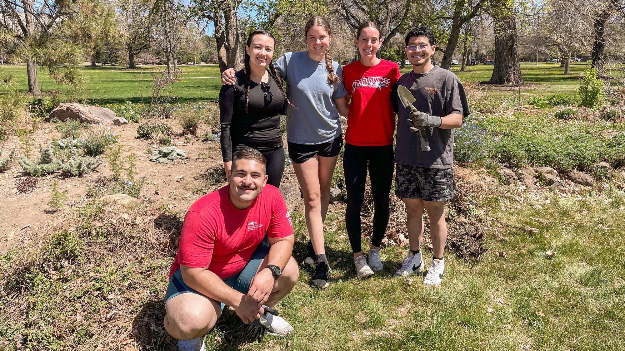 Four students standing and one crouching and holding shovels during a volunteering event
