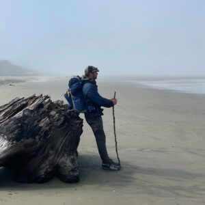 Jonathon Stalls leaning against an old tree on the beach, looking at the Pacific Ocean