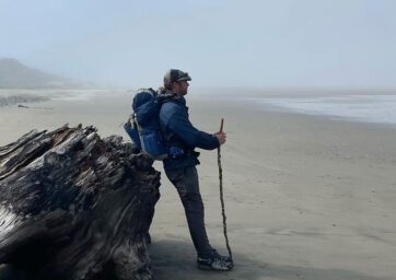 Jonathon Stalls leaning against an old tree on the beach, looking at the Pacific Ocean