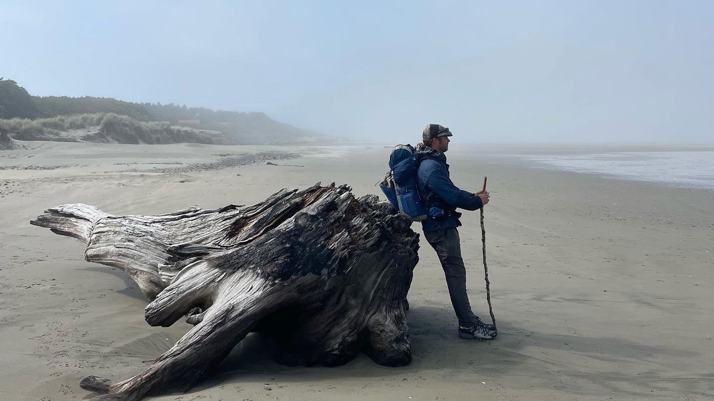 Jonathon Stalls leaning against an old tree on the beach, looking at the Pacific Ocean