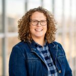 A woman with dark blond, curly hair, wearing green-rimmed glasses.