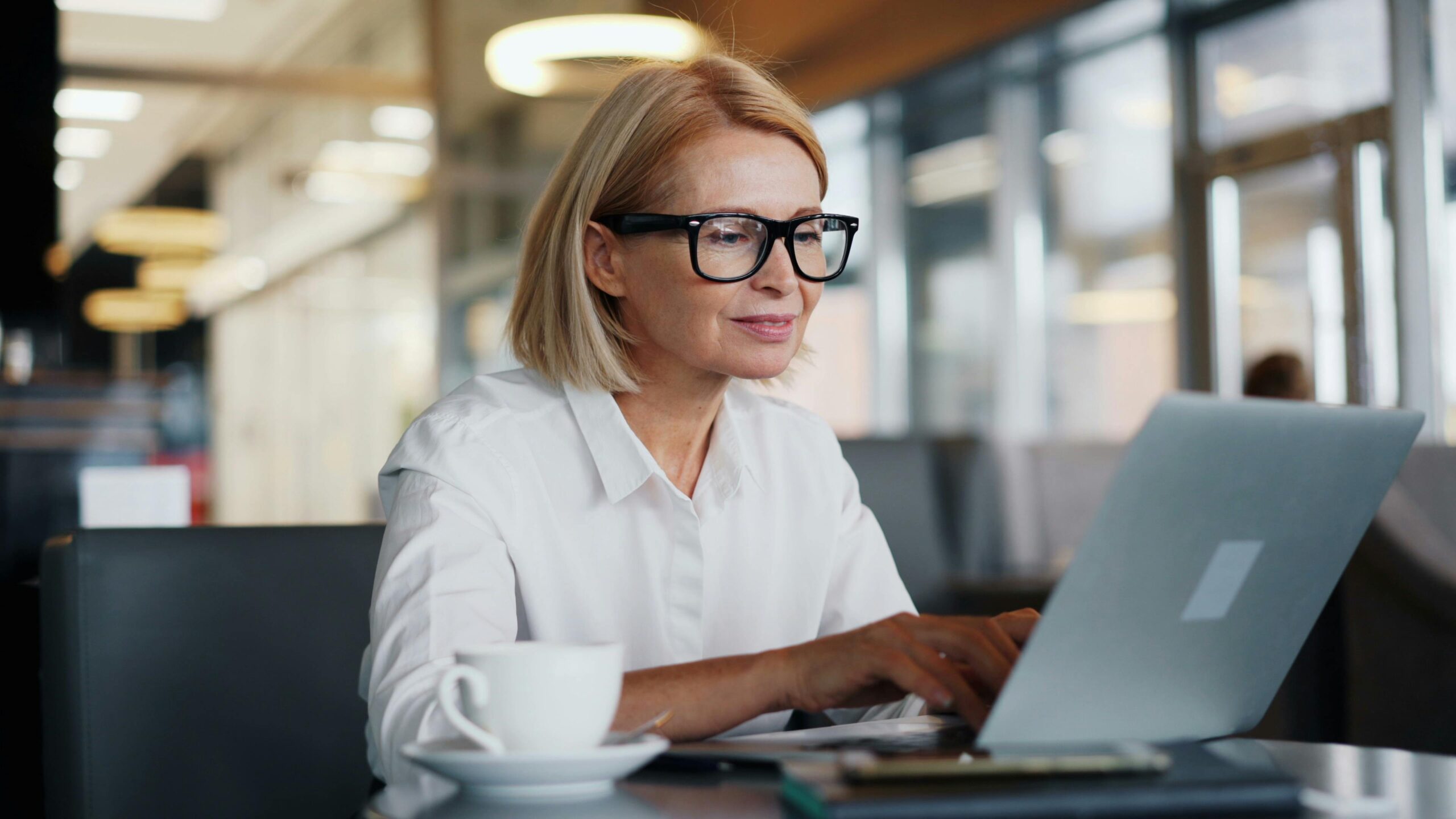 A woman wearing black glasses and a white shirt looks at a computer on a desk.