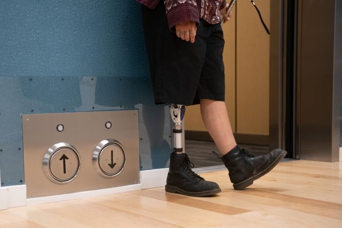 Lower body crop of an Indigenous Two-Spirit person with a prosthetic leg standing next to an open elevator and its floor-located call buttons.