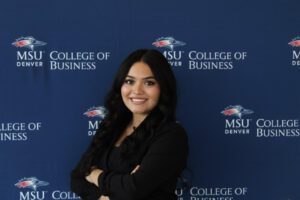 Professional headshot of a smiling woman with dark hair, wearing a black blazer, standing in front of a blue backdrop with the MSU Denver College of Business logo repeated.