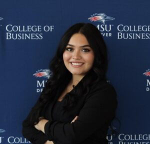 Professional headshot of Victoria with dark hair, wearing a black blazer, standing in front of a blue backdrop with the MSU Denver College of Business logo repeated.