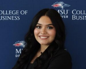 Professional headshot of Victoria with dark hair, wearing a black blazer, standing in front of a blue backdrop with the MSU Denver College of Business logo repeated.