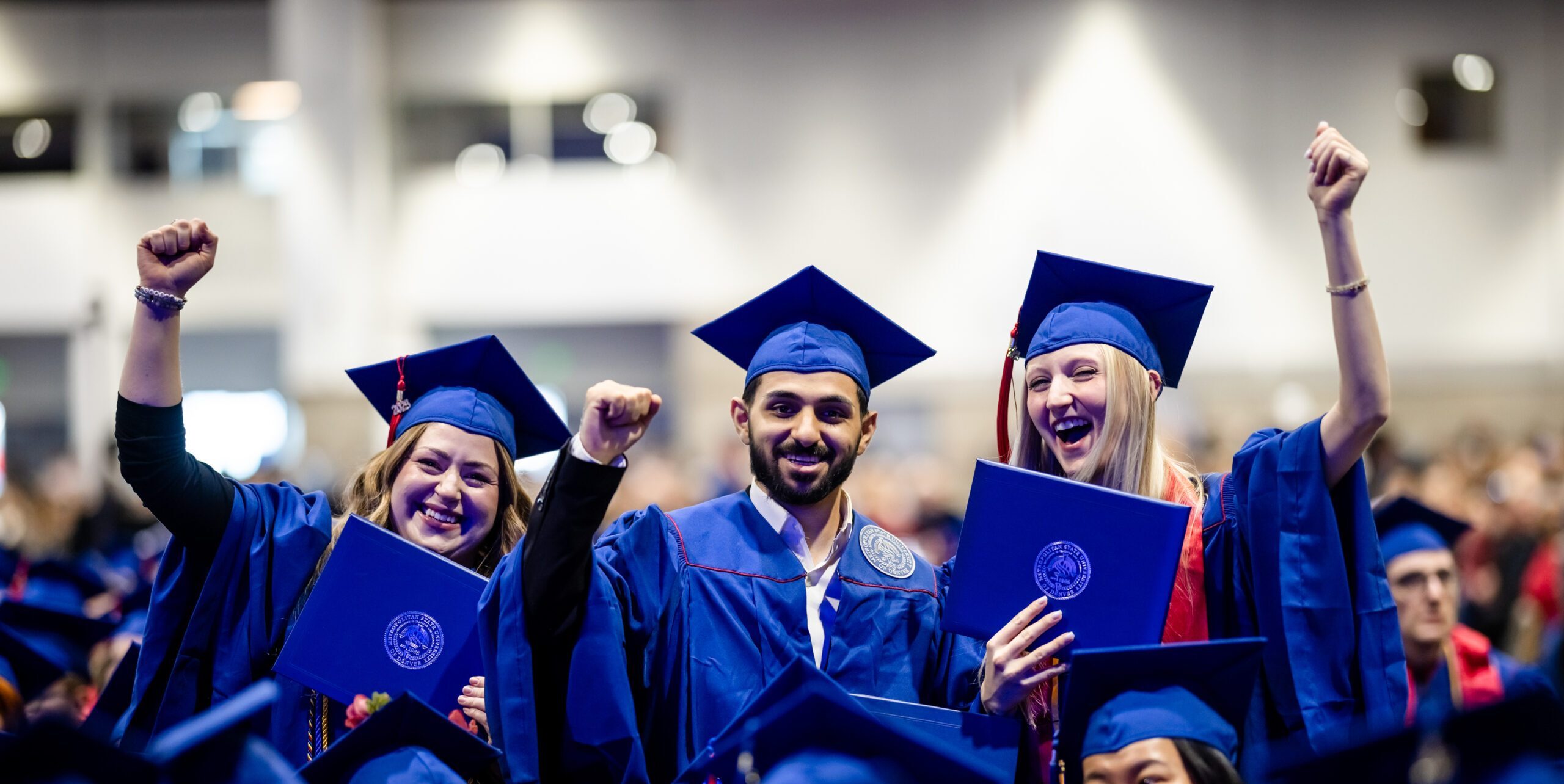 Fall '25 Commencement Ceremony - 3 accounting grads holding up their diploma covers in a sea of graduates