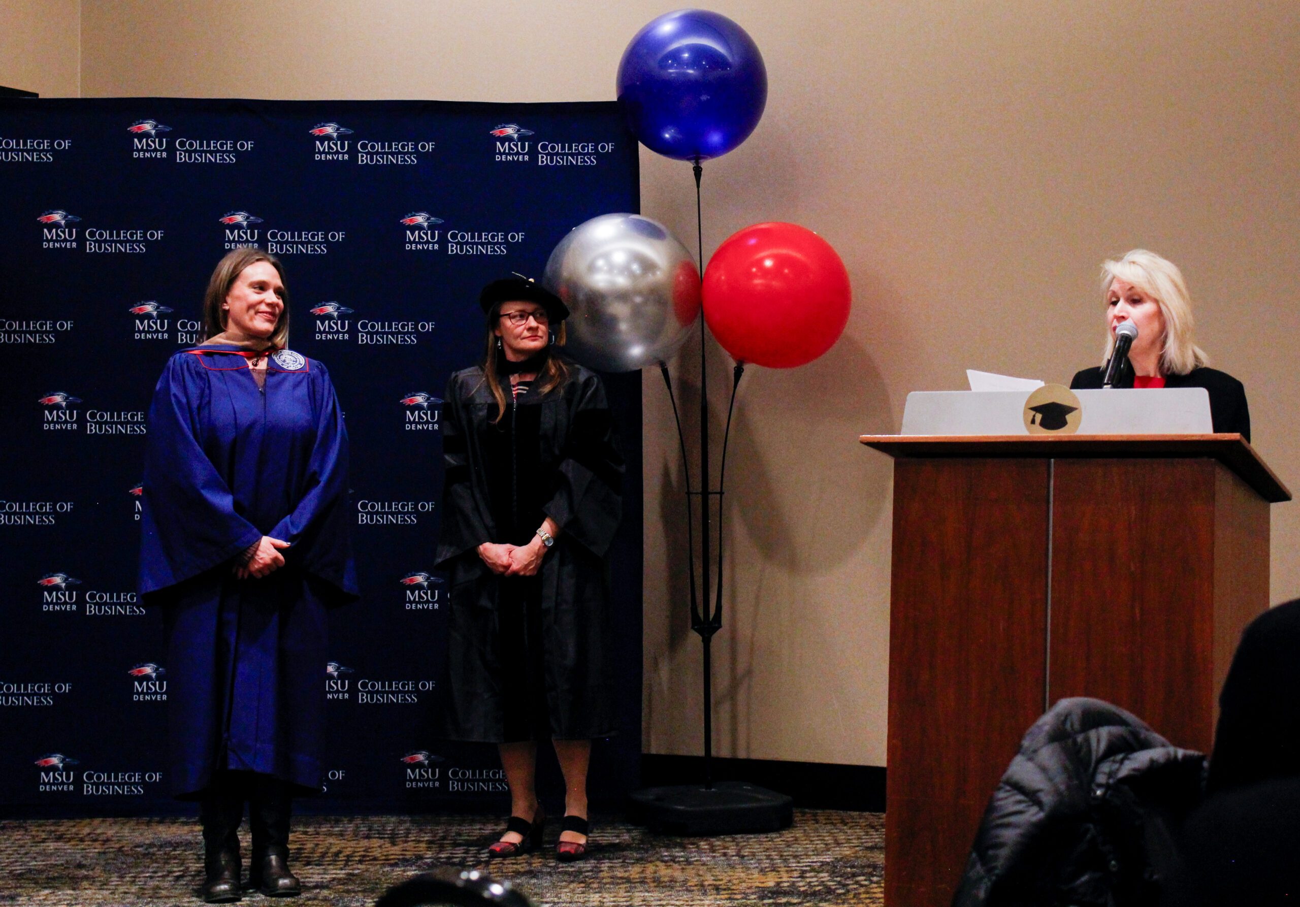 Fall '25 MBA & MPAcc Hooding Celebration - faculty at podium talking about MPAcc grad, Vanessa, after leadership hooded her