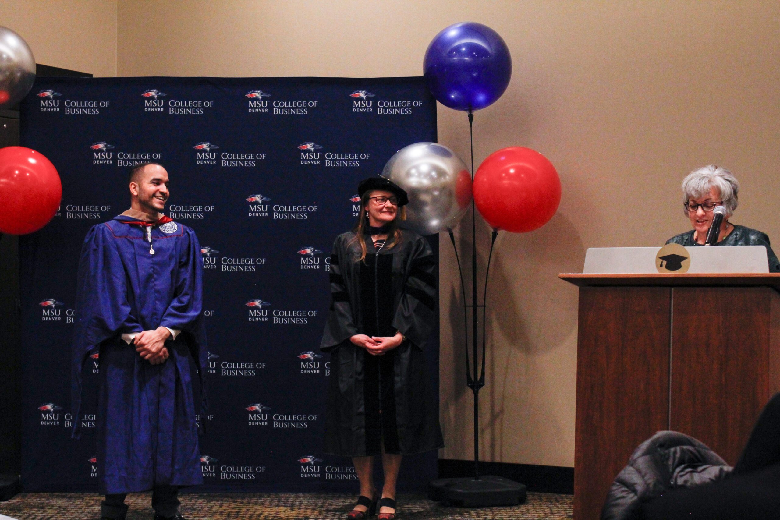 Fall '25 MBA & MPAcc Hooding Celebration - MPAcc faculty, Sheri, at podium talking about MPAcc grad, Kevin as he's being hooded