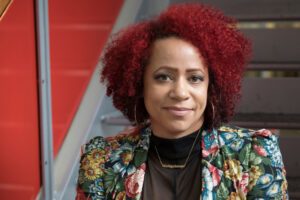 Nikole Hannah-Jones, Black Woman sitting on stairs with Red hair in a floral print Jacket.