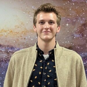 A man with blonde hair wearing a tan jacket stands in front of a wall painted like the galaxy