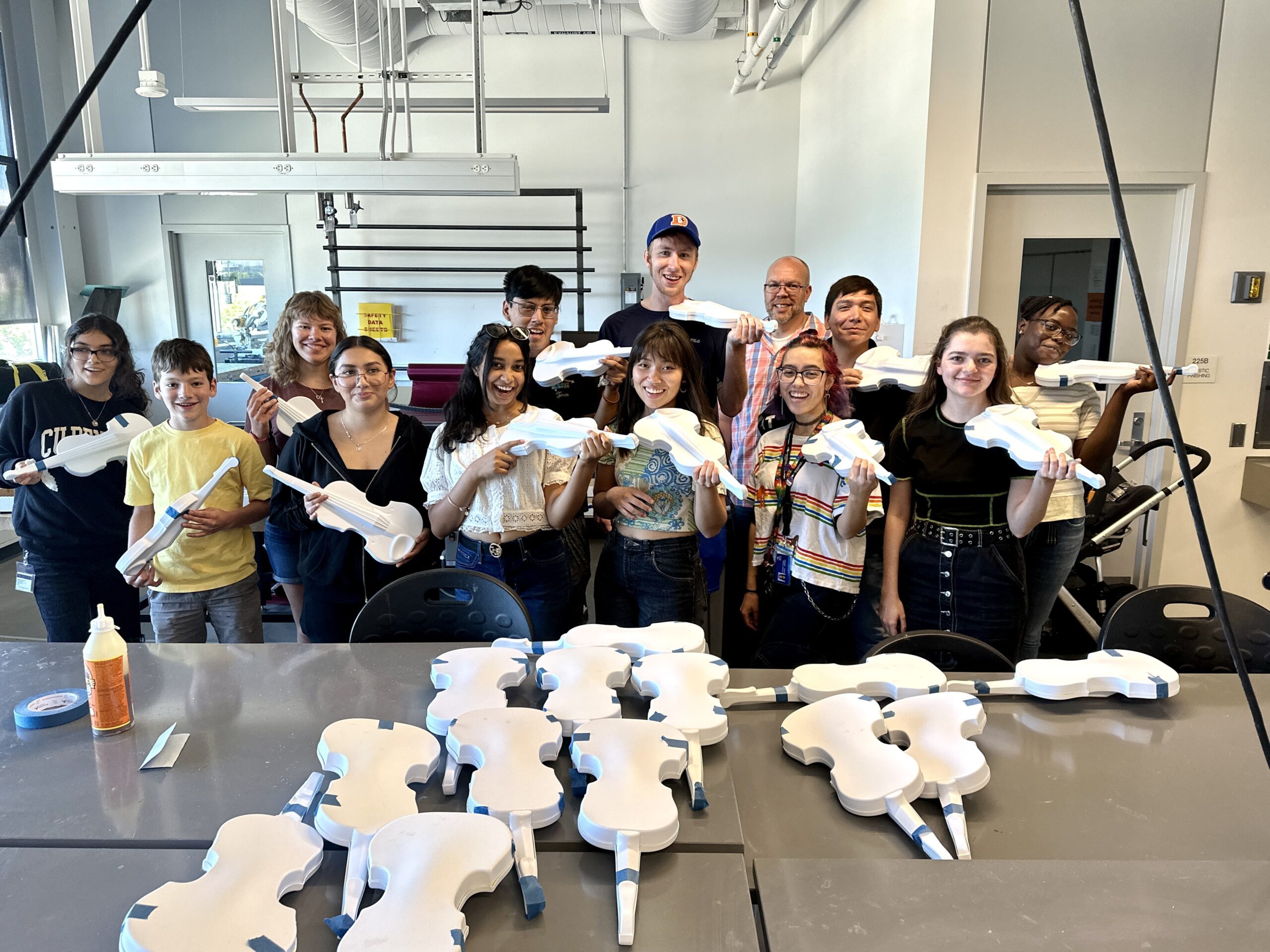 A group of thirteen students and instructors stand in a workshop, smiling and holding white primed violin bodies they have been constructing. The group is arranged in two rows behind a worktable covered with additional white violin bodies in various stages of completion, held together with blue painter's tape. Supplies including wood glue and tape are visible on the table. The setting is an industrial workshop space with exposed ductwork, large windows, and metal shelving.