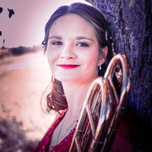 A woman with shoulder length dirty blonde hair holds a trombone and leans against a tree