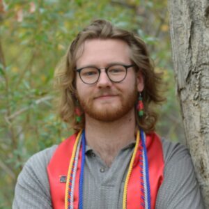 A man with shoulder length dark blonde hair and a beard wearing glasses and graduation cords and a stole