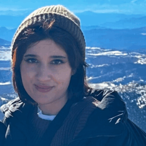 A woman wearing a beanie and winter coat stands in front of a mountain view