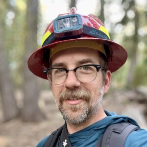 A man with a grey beard and glasses wearing a hard hat with a head lamp