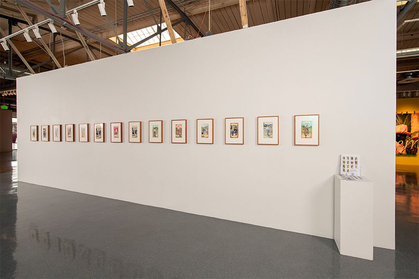 A gallery wall displaying a series of fourteen framed artworks with a stand of brochures on a pedestal.