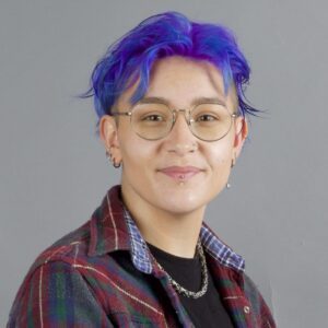 A person with short purple hair and glasses wearing a plaid flannel