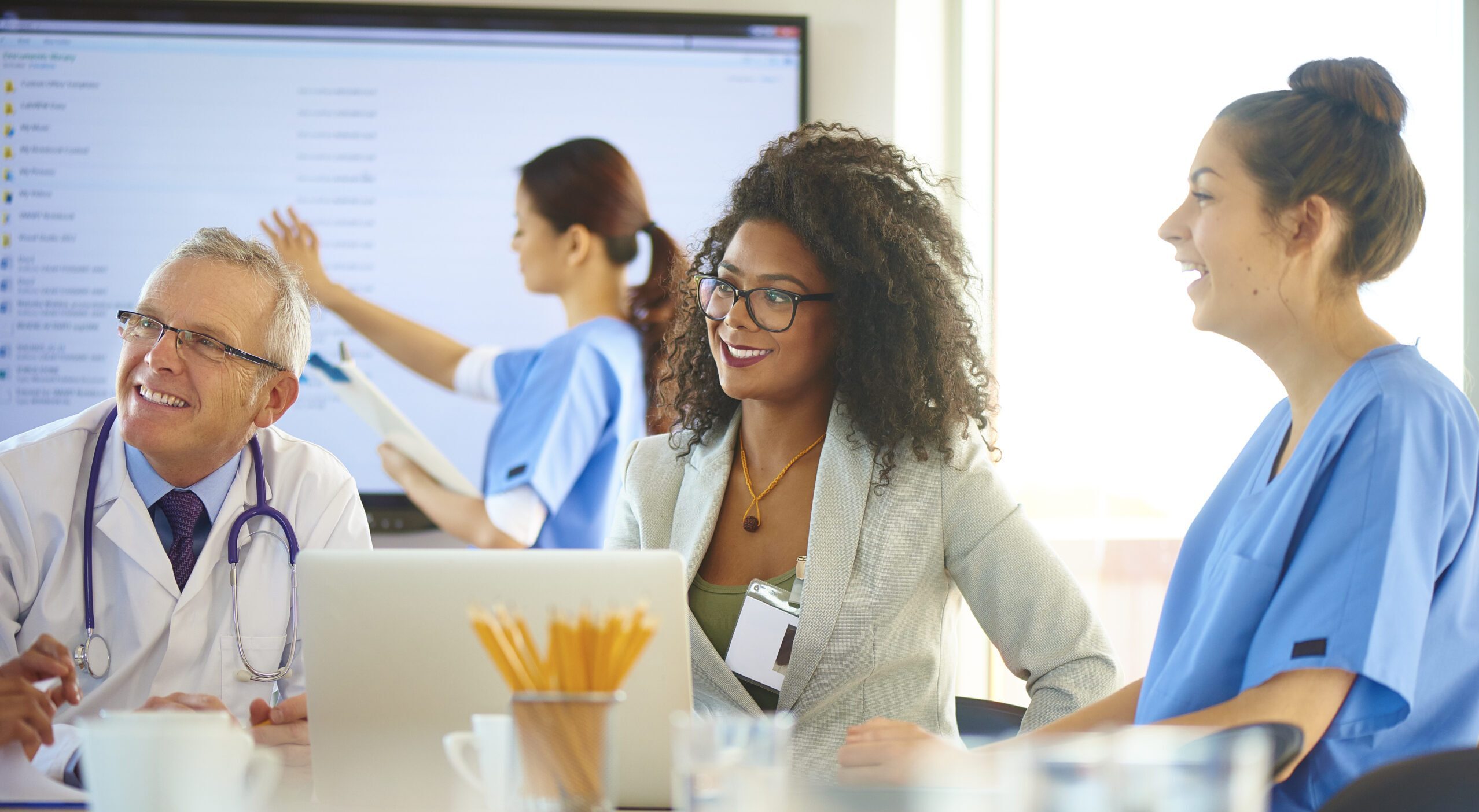 A medical team are discussing something around a boardroom table . They are looking at a laptop and a woman wearing a business suit is listening to the assembled medical team .
