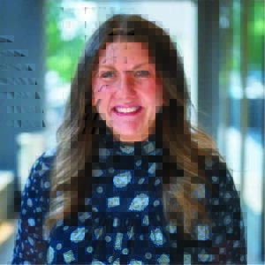 Woman with long brown hair and is wearing a light blue and dark blue spotted shirt.