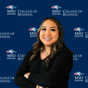 A smiling student ambassador with long, dark wavy hair, wearing a black top and a gold necklace with charms in front of an MSU Denver College of Business backdrop.