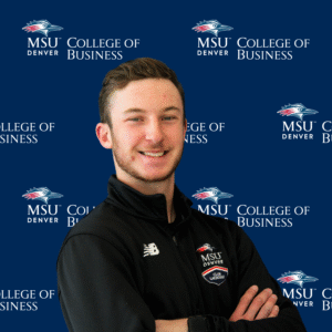 A smiling student ambassador with long, dark wavy hair, wearing a black top and a gold necklace with charms in front of an MSU Denver College of Business backdrop.
