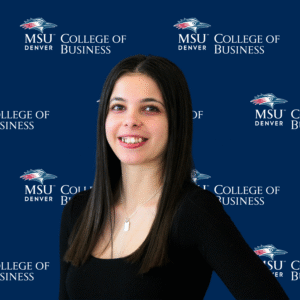 A smiling student ambassador with long, dark wavy hair, wearing a black top and a gold necklace with charms in front of an MSU Denver College of Business backdrop.