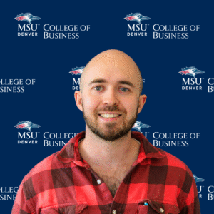 A smiling student ambassador with long, dark wavy hair, wearing a black top and a gold necklace with charms in front of an MSU Denver College of Business backdrop.
