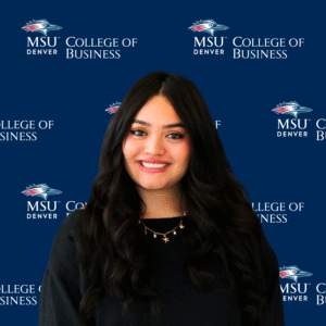 A smiling student ambassador with long, dark wavy hair, wearing a black top and a gold necklace with charms in front of an MSU Denver College of Business backdrop.