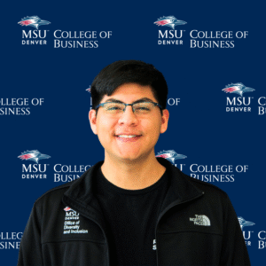 A smiling student ambassador with long, dark wavy hair, wearing a black top and a gold necklace with charms in front of an MSU Denver College of Business backdrop.