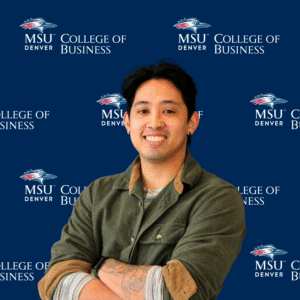 A smiling student ambassador with long, dark wavy hair, wearing a black top and a gold necklace with charms in front of an MSU Denver College of Business backdrop.
