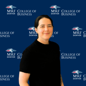 A smiling student ambassador with long, dark wavy hair, wearing a black top and a gold necklace with charms in front of an MSU Denver College of Business backdrop.
