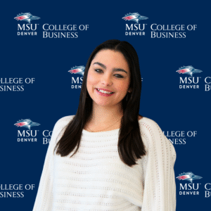A smiling student ambassador with long, dark wavy hair, wearing a black top and a gold necklace with charms in front of an MSU Denver College of Business backdrop.