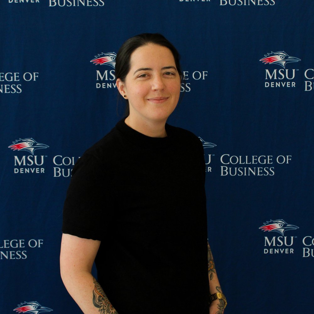 A professional headshot of a student with dark hair tied back, smiling slightly and posing at an angle. They are wearing a black short-sleeved shirt, and colorful tattoos are visible on their forearms. The background is a blue fabric backdrop featuring a repeating 