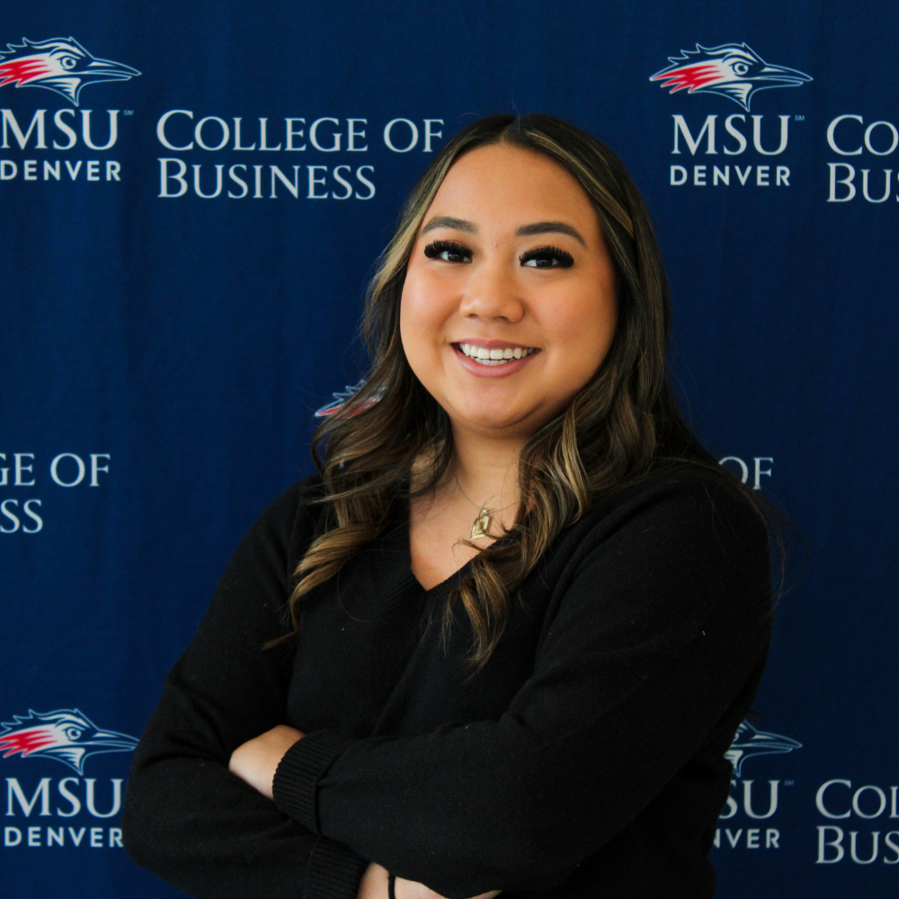 A smiling student ambassador with long, wavy hair with blonde highlights, wearing a black V-neck sweater and crossing her arms in front of an MSU Denver College of Business backdrop.