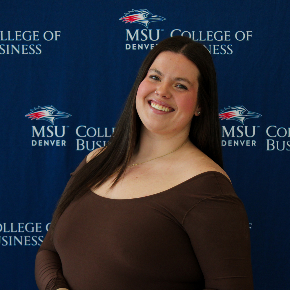 A smiling student ambassador with long brown hair, wearing a brown long-sleeved top and a thin gold necklace in front of an MSU Denver College of Business backdrop.