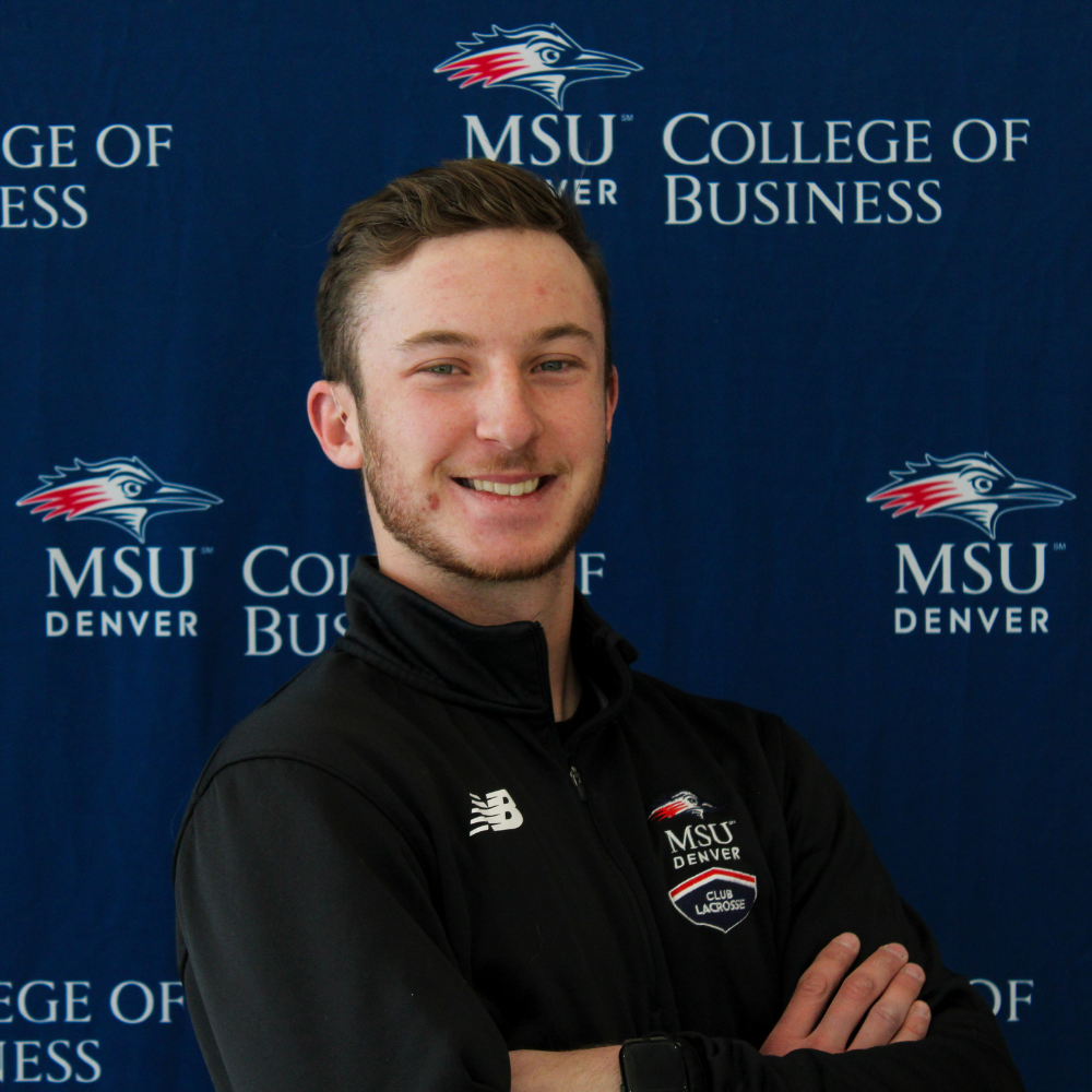 A smiling student ambassador with short brown hair, wearing a black MSU Denver Club Lacrosse zip-up jacket with his arms crossed in front of a College of Business backdrop.
