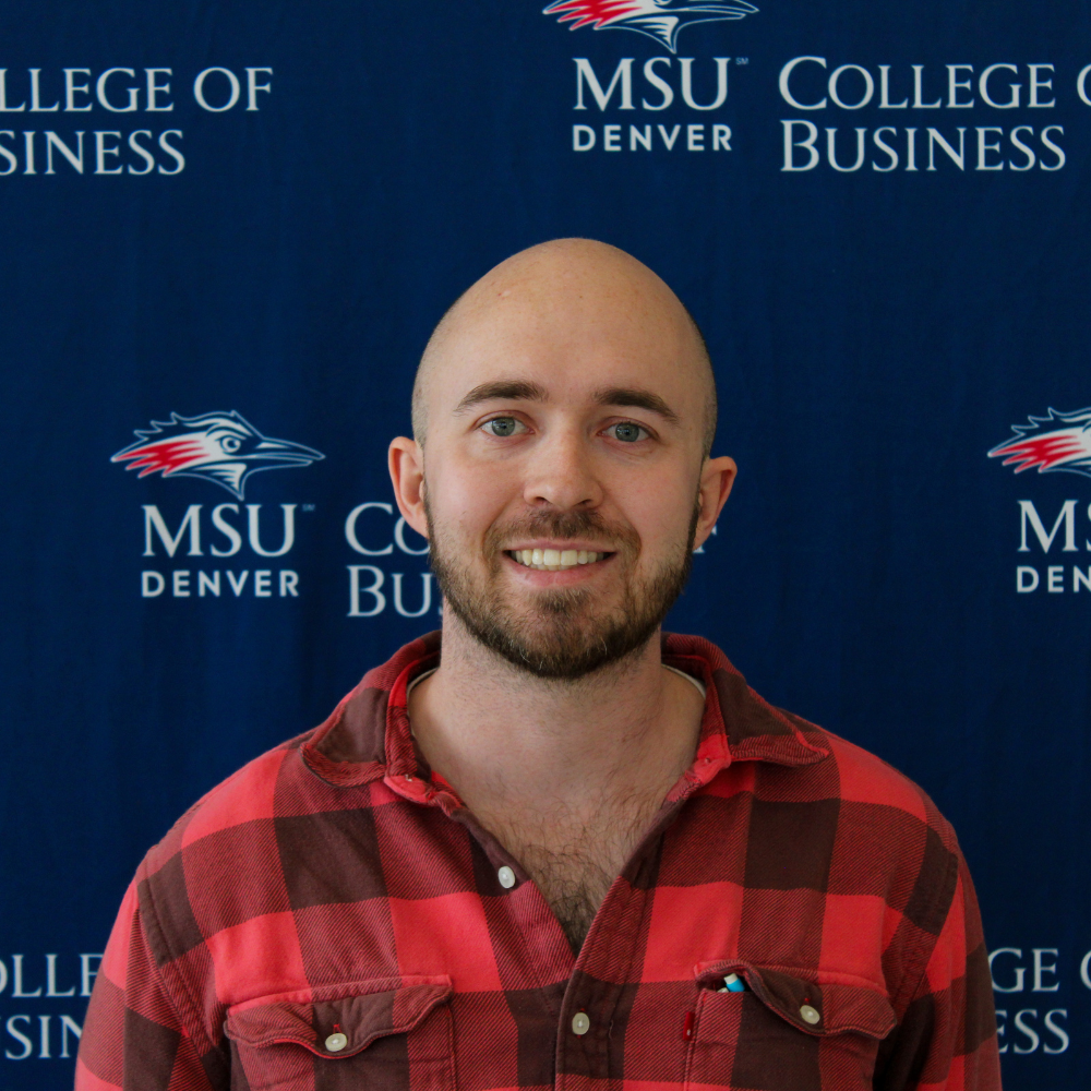 A smiling student ambassador with a beard, wearing a red and black flannel shirt in front of an MSU Denver College of Business backdrop.