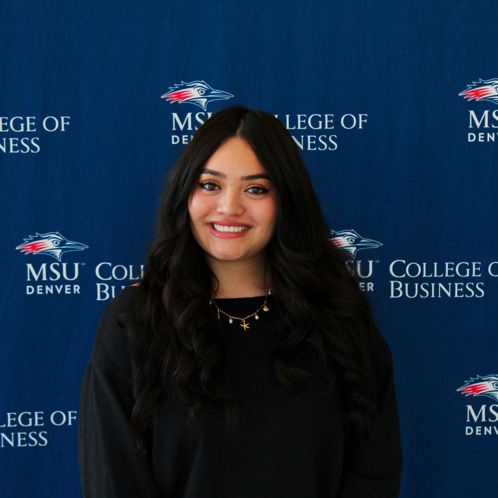 A smiling student ambassador with long, dark wavy hair, wearing a black top and a gold necklace with charms in front of an MSU Denver College of Business backdrop.